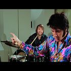 Mann in buntem Hemd vor einem Schlagzeug, zeigt nach unten; weitere Musiker im Hintergrund.