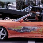 Zwei Sportwagen nebeneinander, ein orangefarbener mit Rennstreifen und ein schwarzer mit freiliegendem Motor. Zwei Fahrer.