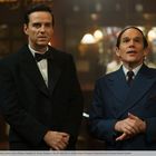 Zwei Männer in eleganter Kleidung, einer im Smoking mit Fliege, der andere im Anzug, stehen nebeneinander in einem edlen Raum.