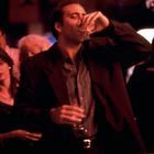 Eine Gruppe von Menschen in einem dunklen, beleuchteten Raum. Eine Person im Vordergrund trinkt aus einem Glas.