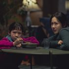 Ein Mädchen in einem pinken Pullover isst an einem Tisch, neben ihr sitzt eine Frau in einem grünen Pullover.