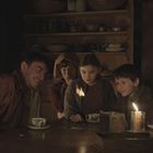 Keine erkennbaren Texte. Eine Gruppe von vier Kindern und einem Erwachsenen sitzt an einem Tisch und betrachtet eine Flamme.