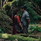 Zwei Menschen stehen sich gegenüber in einem dichten, moosbedeckten Wald. Sie haben eine ernste Unterhaltung.