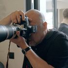 Eine Person mit Kamera fotografiert, während im Hintergrund zwei Menschen stehen, einer mit einer Hundemaske und Lederharnisch.