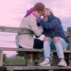 Zwei Personen sitzen auf einer Bank in einer Landschaft bei Sonnenuntergang und küssen sich zärtlich.