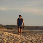 Ich kann keine Texte erkennen. Ein Mann in einem blauen Tanktop geht barfuß am Strand entlang, hält Schuhe in der Hand.