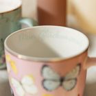 Auf einer Tasse steht "Mein Glücksmoment". Sie ist rosa und mit Schmetterlingen verziert.