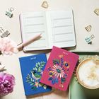 Zwei bestickte Notizbücher mit "2027", ein Kalender, Binder Clips, goldenes Zubehör, Kaffee und Blumen.