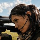 Eine Person zielt mit einem Gewehr, trägt ein Headset und schaut konzentriert. Hintergrund: blauer Himmel mit Wolken.