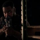 Keine sichtbaren Texte erkennbar. Ein Mann zielt mit einem Gewehr in einem dunklen, möglicherweise industriellen Raum.