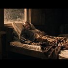 Ein Mann liegt auf einem Bett in einem rustikalen Raum. Sonnenlicht fällt durch ein Fenster.