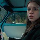 Eine Frau sitzt überrascht in einem blauen Oldtimer. Im Hintergrund sind verschwommene Bäume zu sehen.