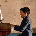 Ein Junge mit Brille spielt konzentriert Klavier in einem schlichten Raum. Er trägt einen dunklen Mantel.