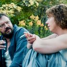 Zwei Personen rauchen und unterhalten sich im Grünen, umgeben von gelbem Laub und Pflanzen.