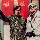 Zwei Männer in Militäruniformen stehen nebeneinander. Im Hintergrund sind rote Fahnen mit gelben Hammer-und-Sichel-Symbolen.