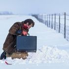 Ein Mann kniet im Schnee, blutverschmiert, mit einem schwarzen Aktenkoffer. Ein Zaun verläuft diagonal im Hintergrund.