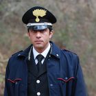 Ein Mann in Uniform mit schwarzem Hut und goldener Verzierung steht vor einem unscharfen, bewaldeten Hintergrund.