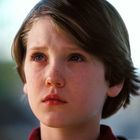 Ein Kind mit Sommersprossen und braunen Haaren blickt nachdenklich in die Ferne, der Hintergrund ist unscharf.