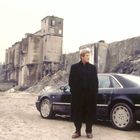 Ein Mann steht vor einem schwarzen Auto, umgeben von verfallenen, industriellen Gebäuden und unebenem Boden.