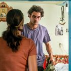 Ein Mann mit lockigem Haar steht einer Frau gegenüber in einem Raum. An der Wand hängen religiöse Bilder und Fotos.