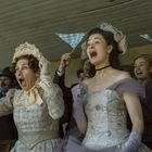 Menschen jubeln begeistert, einige halten blau-weiß-karierte Fahnen. Zwei Frauen vorne tragen historische Kleidung.