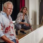 Zwei Personen in blutbefleckten Hemden in einer Halle, eine steht, die andere hockt. Im Hintergrund ein Fenster mit grünem Ausblick.