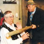 Zwei ältere Männer; einer sitzt mit Bandana, der andere trägt Hut, hält einen Holzstab, beide in traditioneller Kleidung.