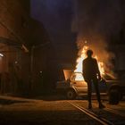 Eine Person steht vor einem brennenden Auto in einer dunklen Gasse, umgeben von Gebäuden und Rauch.