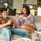 Ein Mann und eine Frau sitzen auf einem Sofa, lächeln und spielen Karten, im Hintergrund eine moderne Küche.