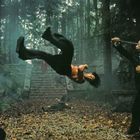 Eine Person führt einen Rückwärtssalto in einem nebligen Wald aus; umgeben von Statuen und kämpfenden Männern.