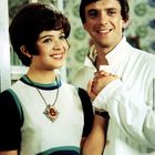 Ein Mann und eine Frau lächeln. Sie trägt ein weißes Kleid mit Schmuckanhänger, er ein weißes Hemd. Sie halten sich an den Händen.