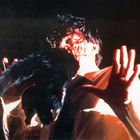 Ein Mensch hält ein haariges Tier vor seinem blutigen Gesicht, Hände ausgestreckt, dramatische Beleuchtung.