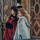 Zwei Frauen in opulenten, historischen Kostümen stehen in einem prunkvollen, palastartigen Raum.