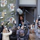 Viele Kameras und Mikrofone umringen einen Mann vor einem Fenster mit Fotos und Notizen.