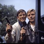 Zwei Männer in Jacken halten lächelnd Pistolen in der Hand; sie scheinen entspannt und stehen im Freien vor einem Baum.