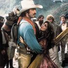 Ein Mann in Cowboy-Kleidung hält eine Frau im Arm. Um sie herum stehen mehrere Männer, teils erstaunt zuschauend.