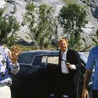 Drei Männer stehen vor einem Auto in einer bergigen Landschaft. Einer hält eine Pistole, ein anderer hebt die Hände.