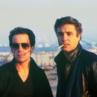 Zwei Männer im Vordergrund, einer mit Sonnenbrille, der andere raucht. Industriehafen im unscharfen Hintergrund.