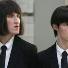 Zwei Personen in schwarzen Anzügen mit Krawatten und dunklen Bob-Frisuren stehen nebeneinander in einem Büro.