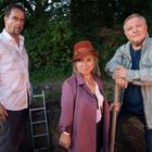 Drei Personen im Freien, umgeben von Bäumen, stehen an einem Erdloch mit Leiter. Eine Person trägt einen rotbraunen Hut.