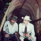 Zwei Männer sitzen nebeneinander in einem hölzernen Wagen. Der eine trägt einen Anzug und Hut, der andere ein kariertes Hemd.