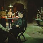 Dunkler Raum mit Labor-Ausstattung. Männer in orientalischer Kleidung arbeiten, andere sitzen, eine Person im Vordergrund sitzt lächelnd.