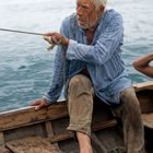 Ein älterer Mann in einem blauen Hemd sitzt in einem Holzboot und hält ein Seil, vor einem ruhigen Ozean.