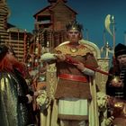 Ein König mit Krone, rotem Zepter und goldenem Gewand, umgeben von Höflingen und Wachen mit Speeren, vor Holzgebäuden.