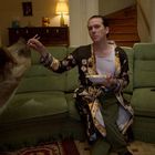 Ein Mann in gemustertem Hausmantel sitzt auf einem grünen Sofa, füttert einen großen Hund mit Essstäbchen.