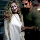 Keine Texte vorhanden. Eine lächelnde Frau in weißem Kleid mit lockigem Haar wird von einem Mann in Raum umarmt.