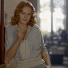 Es gibt keinen Text zu erwähnen. Eine Frau mit lockigem blondem Haar lehnt lässig im Türrahmen, schaut lächelnd nach vorne.