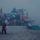 Eine Person zieht einen gelben Mülleimer über einen nebligen Strand, mit einem großen Spielplatz im Hintergrund.