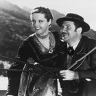 Zwei lachende Personen in traditioneller bayerischer Kleidung, er mit Hut, sie mit geflochtenem Haar. Berge im Hintergrund.