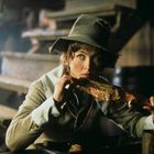 Eine Person mit einem Hut isst ein großes Stück Fleisch in einer rustikalen Umgebung mit Holztischen.